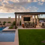 Custom backyard design with slatted metal shade structure, outdoor kitchen with dining area and metal scupper water feature into a blue pool.