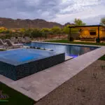 Custom backyard design with natural stone decking, blue raised spa and metal shade structure.