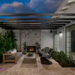 Customized backyard design with slatted metal shade structure, floor-to-ceiling brick fireplace and privacy hedges.