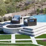 Custom backyard design with metal fencing, circular fire pit and artificial turf pattern.